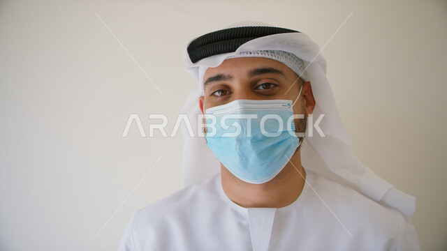 إتباع الاحترازات و التعليمات الوقائيه ضد الفايروسات، تجنب الملامسه السطحيه والحفاظ على المسافات، بورتريه مقرب لرجل عربي خليجي اماراتي يرتدي الكندوره والغتره يضع القناع الطبي الوقائي للوقايه من فايروس كورونا، خلفيه بيضاء.