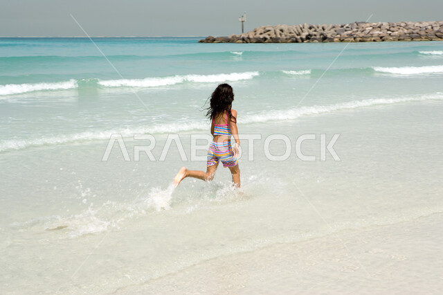 القيام بهواية الركض  في الهواء الطلق على البحر, أنشطة ترفيهية صيفية في العطلة, الإستمتاع بالفعاليات الصيفية الممتعة , فتاة صغيرة عربية إمراتية خليجية تقوم باللعب وتستمتع بالجري على شاطئ البحر, منتجعات شاطئية في الإمارات