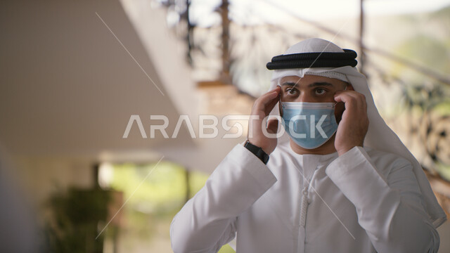 الحمايه من كوفيد-١٩، الحفاظ على السلامة وتجنب العدوى بفايروس كورونا، القيام بالتحضيرات والاستعداد للخروج من المنزل، صورة مقربه لرجل عربي خليجي اماراتي يقوم بوضع الكمامة ويرتدي الكندورة والغترة  الاماراتية .