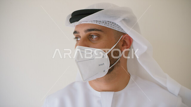 إتباع الاحترازات  والتعليمات الوقائيه ضد كوفيد 19 ، بورتريه مقرب لرجل عربي خليجي اماراتي يرتدي الكندوره والغتره يضع القناع الطبي الوقائي للوقايه من فايروس كورونا وينظر إلى شيء ما، الحفاظ على المسافات وتجنب الملامسه السطحيه، خلفيه بيضاء.