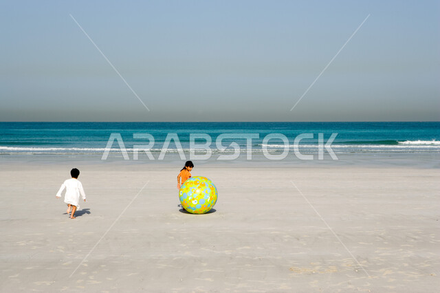 أخوان يقومان بقضاء الإجازة الصيفية باللعب معا في أجواء صيفية ممتعة, فتاة صغيرة عربية إمراتية خليجية تقوم باللعب على شاطئ البحر بالكرة المطاطية الكبيرة رفقة أخيها في رحلة صيفية, أنشطة ترفيهية عائلية في العطلة, منتجعات شاطئية في الإمارات