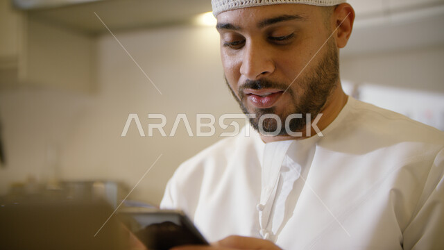 متابعه الأخبار الجديدة على وسائل الاعلام بإيماءات السعادة، إرسال الرسائل النصية والتواصل مع الأصدقاء والأقارب في وقت النهار, صورة مقربه لشاب عربي إماراتي خليجي مبتسم يرتدي الكندورة والطاقية البيضاء يجلس على الأريكة ويحمل بيده الهاتف المحمول