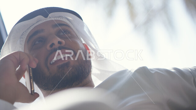 تبادل الأحاديث المسلية مع الأصدقاء والأقارب، إجراء مكالمة هاتفية صوتية عبر الجوال، ايماءات تدل على السعادة، صورة مقربة لرجل عربي اماراتي خليجي يرتدي الكندورة والغترة الاماراتية يجلس في غرفة المعيشة ويحمل بيده الهاتف المحمول