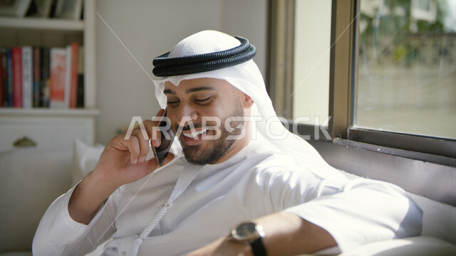 إجراء مكالمة هاتفية صوتية عبر الجوال، ايماءات تدل على السعادة، صورة مقربة لرجل عربي اماراتي خليجي يرتدي الكندورة والغترة الاماراتية يجلس في غرفة المعيشة ويحمل بيده الهاتف المحمول، الضحك والتسامر مع الأصدقاء والأقارب