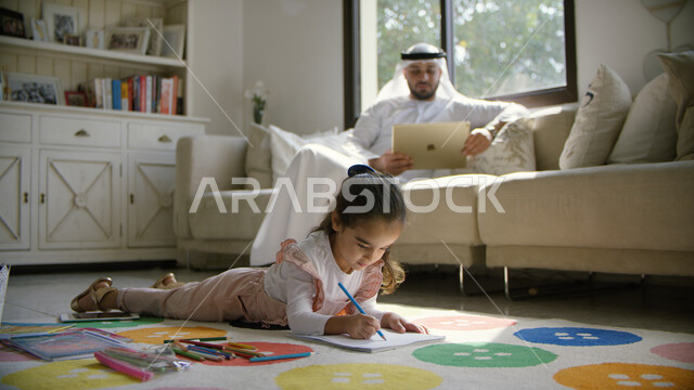 الألعاب المسلية و التعليمية عبر الانترنت مع الأبناء، الترابط الأسري والمشاركة مع الأولاد النشاطات الترفيهيه، استخدام أجهزة التابليت في المنزل والاستفادة من أوقات الفراغ, والد عربي خليجي إماراتي يرتدي الكندورة والغترة يجلس في غرفة المعيشة برفقه ابنته