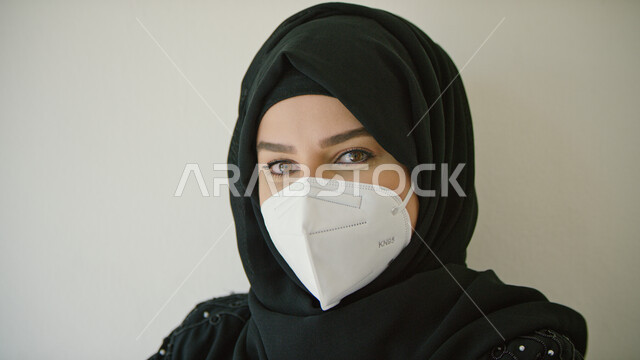 النظر إلى الكاميرا، التقيد بقوانين وقواعد وزاره الصحة لتجنب الاصابه بكوفيد-١٩، بورتريه مقرب لامرأة عربية خليجية اماراتية محجبة ترتدي عباءه سوداء وتضع الكمامة لتجنب تفشي فايروس كورونا، خلفية بيضاء.