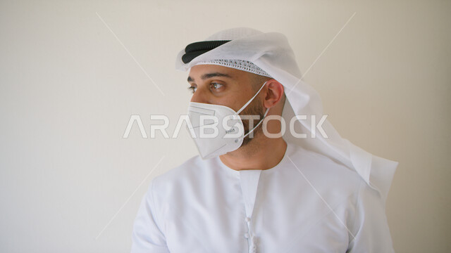 إتباع الإجراءات و الاحترازات الوقائيه ضد كوفيد 19 ، بورتريه مقرب لرجل عربي خليجي اماراتي يرتدي الكندوره والغتره يضع القناع الطبي الوقائي للوقايه من فايروس كورونا وينظر إلى شيء ما، الحفاظ على المسافات وتجنب الملامسه السطحيه، خلفيه بيضاء.