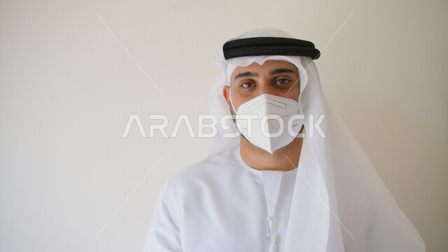 إتباع الإجراءات الوقائيه ضد الفايروسات، النظر إلى الكاميرا، تجنب الملامسه السطحيه والحفاظ على المسافات، بورتريه مقرب لرجل عربي خليجي اماراتي يرتدي الكندوره والغتره يضع القناع الطبي الوقائي للوقايه من فايروس كورونا، الاهتمام بالمظهر الخارجي، خلفيه بيضاء.