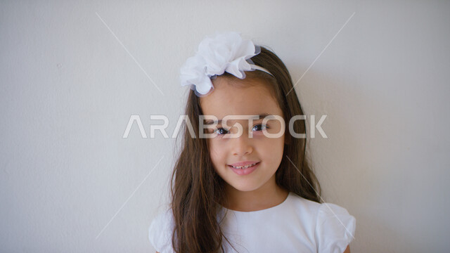 ايماءات تدل على السعادة، التقاط صور شخصية للوثائق المدرسية, الإهتمام بالمظهر الخارجي للأطفال الصغار، بورتريه مقرب لطفلة عربيه خليجيه إماراتية تنظر إلى الكاميرا مبتسمة ترتدي فستان أبيض اللون، ارتداء الملابس الجديده في الأعياد والمناسبات، خلفيه بيضاء