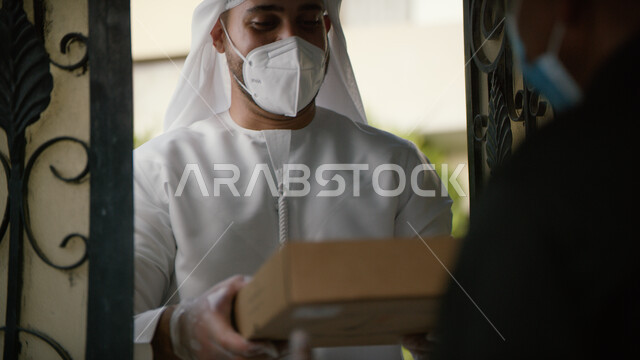 خدمة توصيل طلبيات ومشتريات الاونلاين الى المنزل، صورة من الخلف لمندوب التوصيل يقوم بتسليم صندوق الطلبية لرجل عربي خليجي اماراتي يرتدي الكندورة والغترة الاماراتية الالتزام بالاجراءات الاحترازية وارتداء الكمامة والكفوف للوقاية من الامراض وفايروس كورونا.