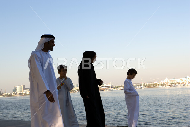 The concept of family bonding and spending time walking with children, taking care of the family and sharing happy moments with them, an Emirati Arab family strolling and walking on the Corniche on the day off, family dialogues and discussions abroad