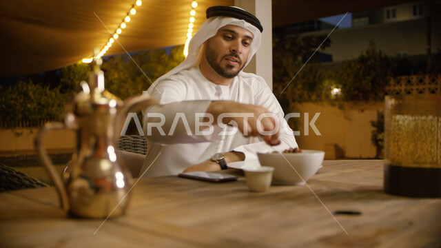 تناول حبات التمر المكنوز الاماراتي, زينة و أضواء الشهر الفضيل, وجبه صحيه ذات قيمه غذائيه عالية، صورة مقربه لرجل عربي إماراتي خليجي يرتدي الغترة والكندورة الاماراتية يحمل بيده يتناول وجبة الفطور في إحدى المطاعم