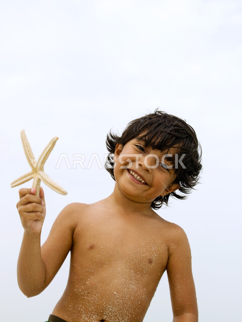 النظر إلى الكاميرا , صورة مقربة لصبي عربي إماراتي خليجي مبتسم يلعب ينجم البحر على الشاطئ, قضاء أوقات ممتعة ومسلية في العطلة, السعادة والاستمتاع بالفعاليات الصيفية، أماكن سياحية ترفيهية في الخليج العربي، اكتشاف الأولاد الصدف والقذائف البحرية.