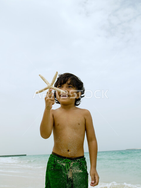  اكتشاف الأولاد الصدف والقذائف البحرية، صورة مقربة لصبي عربي إماراتي خليجي مبتسم يلعب ينجم البحر على الشاطئ, قضاء أوقات ممتعة ومسلية في العطلة, إيماءات وجه تدل على السعادة والاستمتاع بالفعاليات الصيفية، أماكن سياحية ترفيهية في الخليج العربي.