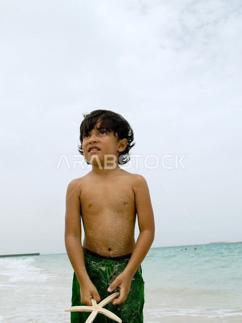  اكتشاف الأولاد الصدف والقذائف البحرية، صورة مقربة لصبي عربي إماراتي خليجي يلعب ينجم البحر على الشاطئ, قضاء أوقات ممتعة ومسلية في العطلة, السعادة والاستمتاع بالفعاليات الصيفية، أماكن سياحية ترفيهية في الخليج العربي.