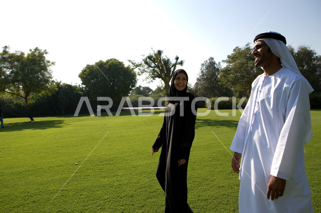 الحوارات والنقاشات العائلية في الهواء الطلق, منظر العشب والأشجار الخضراء في وضح النهار، زوجان عربيان خليجيان إماراتيان يقومان بالمشي والتنزه في الحديقة العامة في الإمارات، مفهوم الرابطة الزوجية والألفة بين الزوجين