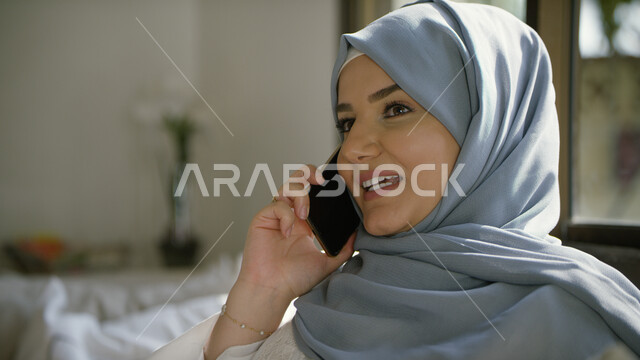 Enjoying talking and entertaining through smart devices and completing work tasks, using modern technologies, a close-up of a smiling, veiled Arab Emirati woman making a phone call via mobile, social communication and chatting with friends with gestures o