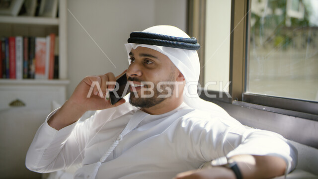 ايماءات تدل على السعادة، إجراء مكالمة هاتفية صوتية عبر الجوال، صورة مقربة لرجل عربي اماراتي خليجي يرتدي الكندورة والغترة الاماراتية يجلس في غرفة المعيشة ويحمل بيده الهاتف المحمول، التواصل مع الأصدقاء والأقارب