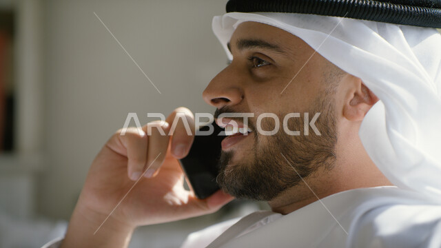 مفهوم صلة الرحم، ايماءات تدل على السعادة، إجراء مكالمة هاتفية صوتية عبر الجوال، صورة مقربة لرجل عربي اماراتي خليجي يرتدي الكندورة والغترة الاماراتية يجلس في غرفة المعيشة ويحمل بيده الهاتف المحمول، التواصل مع الأصدقاء والأقارب