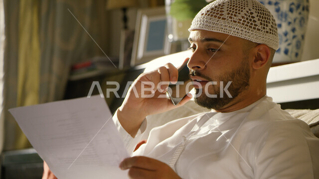 مفهوم ادارة الأعمال وعقد الاجتماعات عبر الانترنت، إنجاز مهام العمل في المنزل، صورة مقربة لرجل عربي خليجي إماراتي يرتدي الكندورة والطاقية الاماراتية البيضاء يقوم بإجراء مكالمة عبر الجوال، العمل عن بعد في زمن الكورونا واتباع طرق الوقاية