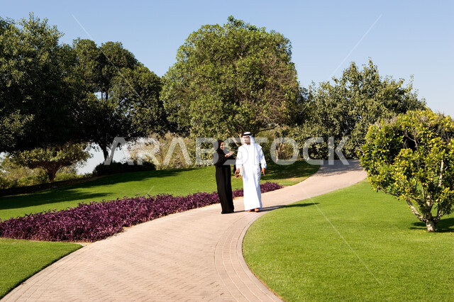 الحوارات والنقاشات العائلية في الهواء الطلق, منظر العشب والأشجار الخضراء في وضح النهار، زوجان عربيان خليجيان إماراتيان يقومان بالمشي والتنزه في الحديقة العامة في الإمارات، مفهوم الرابطة الزوجية والألفة بين الزوجين