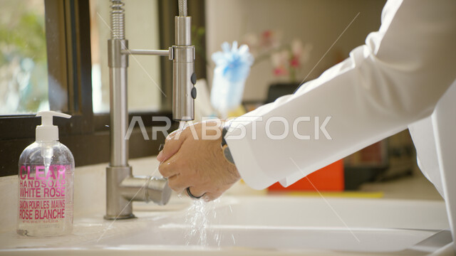 تجنب إنتشار العدوى و اتباع اساليب الحرص، مفهوم العناية بالنظافة الشخصيه، صورة مقربة ليدين رجل عربي خليجي اماراتي يرتدي الكندوره يقوم بغسل يديه بالماء والصابون، ضروره الإلتزام بالتعليمات واجراءات الحماية من فايروس كورونا.