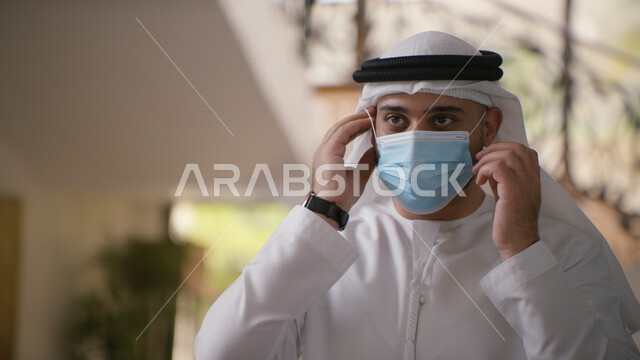 القيام بالتحضيرات والاستعداد للخروج من المنزل، الحمايه من كوفيد-١٩، الحفاظ على السلامة وتجنب العدوى بفايرو كورونا، صورة مقربه لرجل عربي خليجي اماراتي يقوم بوضع الكمامة ويرتدي الكندورة والغترة الاماراتية .