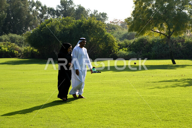 إجراء المحادثات العائلية في الهواء الطلق, منظر العشب والأشجار الخضراء في وضح النهار، زوجان عربيان خليجيان إماراتيان يقومان بالمشي والتنزه في الحديقة العامة في الإمارات، مفهوم الرابطة الزوجية والألفة بين الزوجين