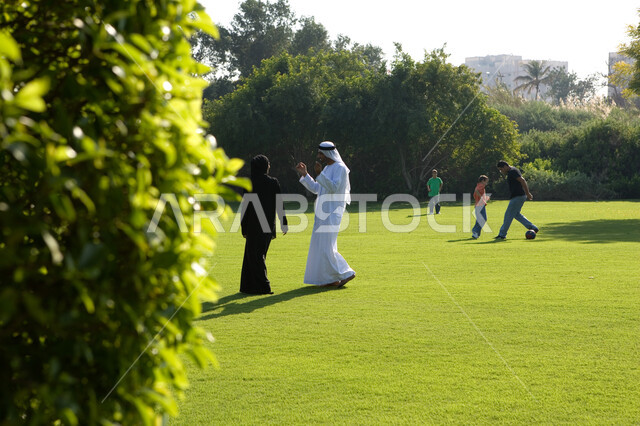 منظر العشب والأشجار الخضراء في الصيف، الحوارات والنقاشات العائلية في الهواء الطلق, صورة من الخلف لزوجان عربيان خليجيان إماراتيان يقومان بالمشي والتنزه في الحديقة العامة، أطفال يلعبون في المساحات الخضراء الواسعة, مفهوم الرابطة الزوجية والألفة بين الزوجين