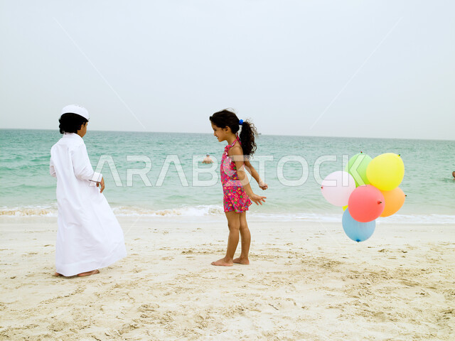 فعاليات ترفيهية مسلية للأطفال, أخ وأخت يلعبان على الشاطئ بالبلالين الملونة, قضاء يوم العطلة على البحر, السياحة في الإمارات العربية، أنشطة ترفيهية أوقات ممتعة.