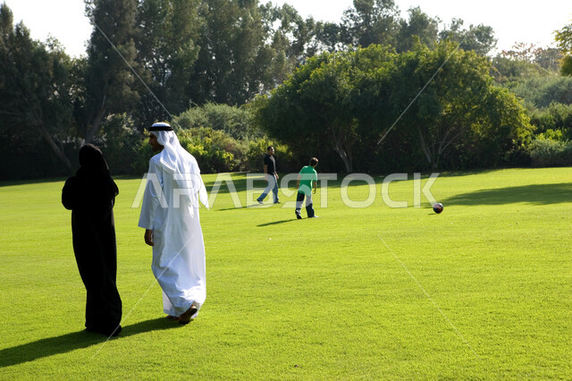 Landscape of grass and green trees in the summer, family dialogues and discussions in the open air, a back image of an Arab Emirati couple walking and hiking in the public garden, children playing in the vast green spaces, the concept of marital bond and 