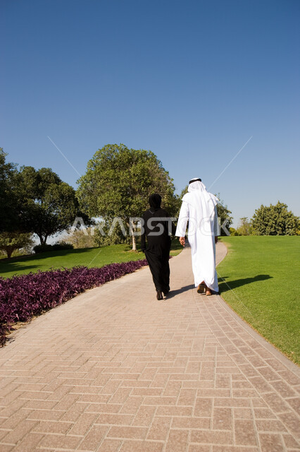 التنزه والسير في الهواء الطلق, منظر العشب والأشجار الخضراء في وضح النهار، صورة من الخلف زوجان عربيان خليجيان إماراتيان يقومان بالمشي والتنزه في الحديقة العامة في الإمارات، مفهوم الرابطة الزوجية والألفة بين الزوجين