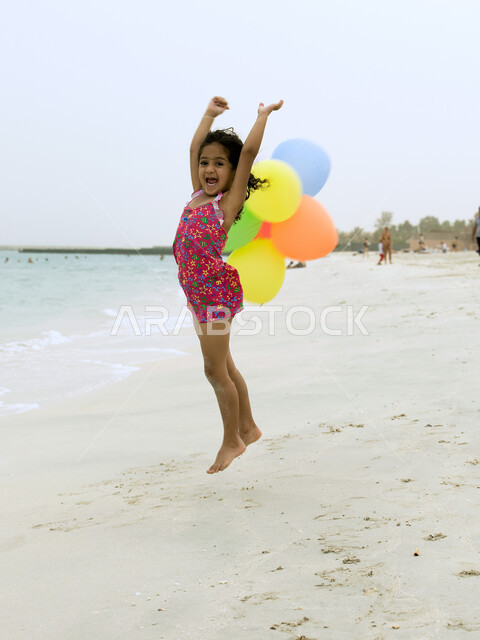 Face and body gestures indicating happiness and enjoyment, picnics on vacation at the sea, entertaining activities for children, an Arab Emirati girl wearing a swimsuit jumping high with joy, playing on the beach with colored balloons, tourism in the UAE,
