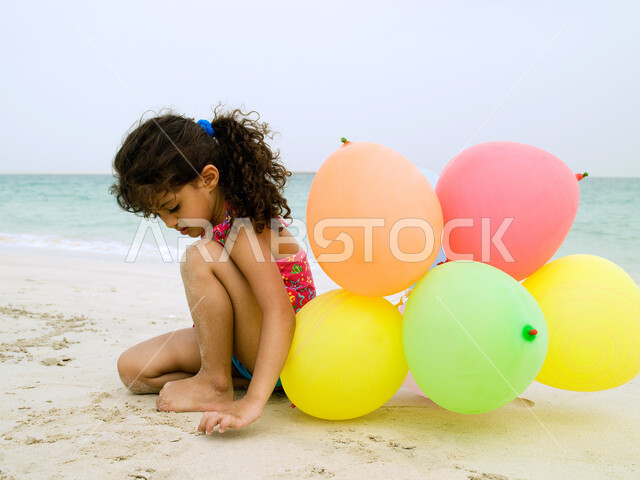 التنزه يوم العطلة على البحر, فعاليات ترفيهية مسلية للأطفال, طفلة عربية خليجية إماراتية ترتدي ملابس السباحة وتلعب على الشاطئ بالرمال الذهبية الناعمة وبالبلالين, السياحة في الإمارات العربية، أنشطة ترفيهية وقضاء أوقات ممتعة.