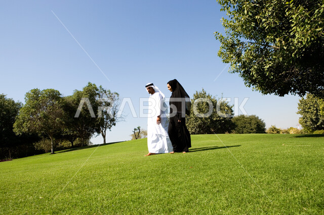 Spending fun times in public parks, having family conversations in the open air, the view of grass and green trees in broad daylight, an Emirati Arab couple walking and hiking in a public park in the Emirates, the concept of marital bond and intimacy betw