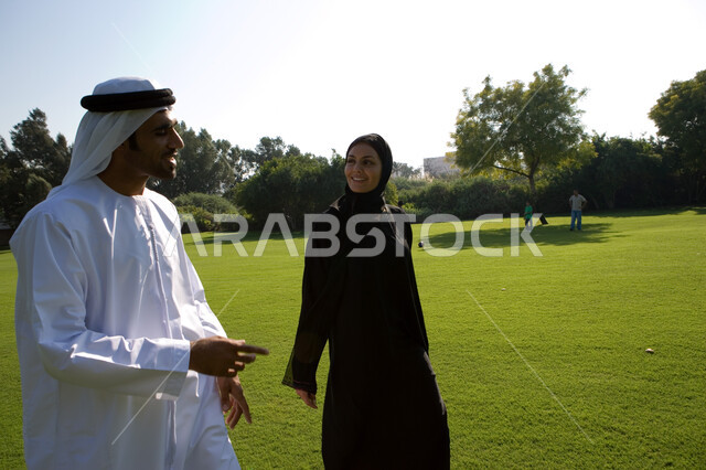 ايماءات تدل على السعادة والاستمتاع بالتحدث, إجراء المحادثات العائلية في الهواء الطلق, منظر العشب والأشجار الخضراء في وضح النهار، صورة من الجانب لزوجين عربين خليجين إماراتيين يقومان بالمشي والتنزه في الحديقة، مفهوم الرابطة الزوجية والألفة بين الزوجين