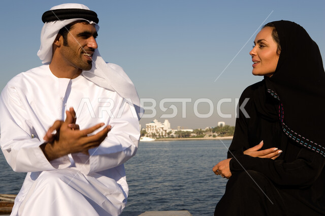 الإنصات والاستماع باحترام للحديث بين الزوجين, قضاء وقت ممتع في جلسات عائلية على الكورنيش في وضح النهار، صورة مقربة لزوجين عربيين خليجيين إماراتيين يقومان بتبادل الأحاديث أثناء الجلوس على الكورنيش بإيماءات الحب و التفاهم، السياحة الساحلية في الامارات