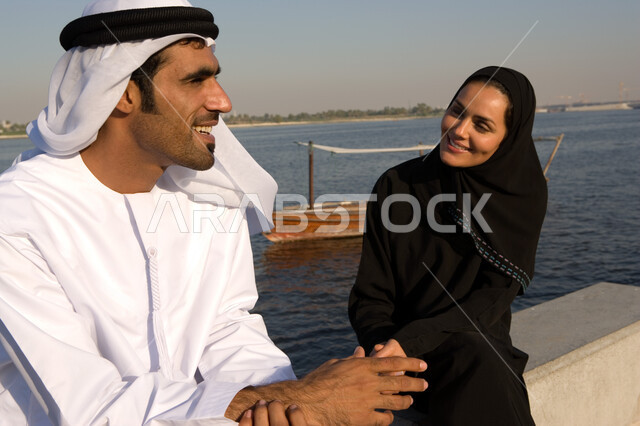 الإنصات والاستماع باحترام للحديث بين الزوجين, قضاء وقت ممتع في جلسات عائلية على الكورنيش في وضح النهار، صورة مقربة لزوجين عربيين خليجيين إماراتيين يقومان بتبادل الأحاديث أثناء الجلوس على الكورنيش بإيماءات الحب و التفاهم، السياحة الساحلية في الامارات