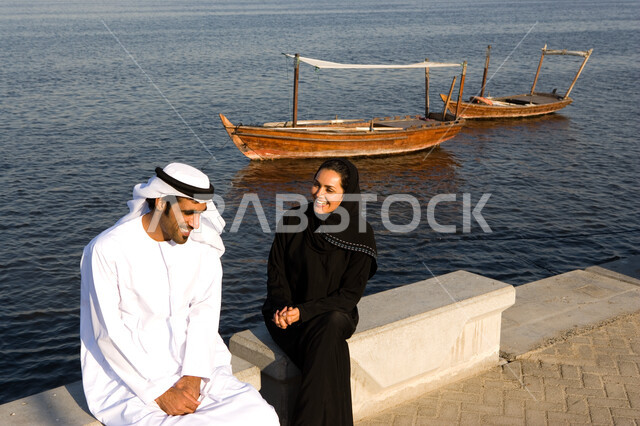 قضاء وقت ممتع في جلسات عائلية على الكورنيش في وضح النهار، زوجان عربيان خليجيان إماراتيان يقومان بتبادل الأحاديث أثناء الجلوس على الكورنيش بإيماءات الحب و التفاهم، السياحة الساحلية في الامارات, الإنصات والاستماع باحترام للحديث بين الزوجين