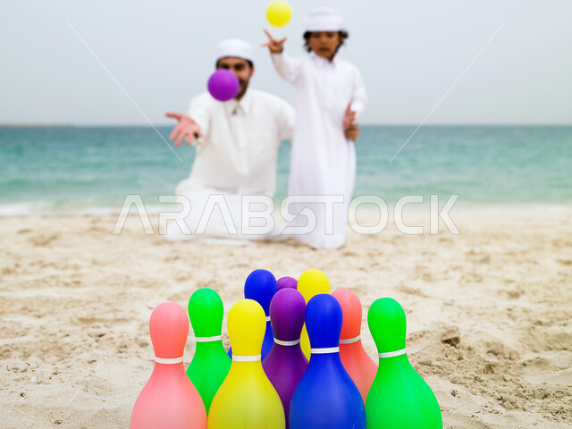 تعلم التركيز وإصابة الهدف، مشاركة الآباء الأبناء نشاطاتهم الترفيهية, قضاء أوقات صيفية ممتعة, صبي صغير عربي خليجي اماراتي يرتدي الزي الإماراتي التقليدي يقوم باللعب بلعبة البولينج رفقة والده على شاطئ البحر، السياحة في الإمارات. 