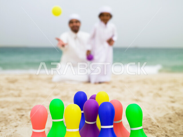 Learning to focus and hit the target, parents sharing their recreational activities with children, spending fun summer times, a young Emirati Arab Gulf boy dressed in traditional Emirati dress playing bowling with his father on the seashore, tourism in th