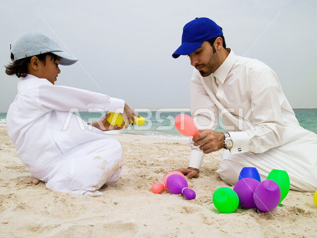 تعلم التركيز وإصابة الهدف، مشاركة الآباء الأبناء نشاطاتهم الترفيهية, قضاء أوقات صيفية ممتعة, صبي صغير عربي خليجي اماراتي يرتدي الزي الإماراتي التقليدي يقوم باللعب بلعبة البولينج رفقة والده على شاطئ البحر، السياحة في الإمارات. 