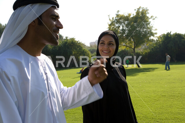 ايماءات تدل على السعادة والاستمتاع بالتحدث, إجراء المحادثات العائلية في الهواء الطلق, منظر العشب والأشجار الخضراء في وضح النهار، صورة مقربة من الجانب لزوجين عربين خليجين إماراتيين يقومان بالمشي والتنزه في الحديقة، مفهوم الرابطة الزوجية والألفة بين الزوجين