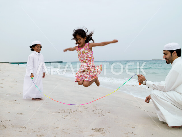 مشاركة الآباء الأبناء نشاطاتهم الترفيهية, قضاء أوقات عائلية ممتعة في الإجازة الصيفية, رجل عربي خليجي اماراتي يرتدي الزي الإماراتي التقليدي يقوم باللعب مع أولاده بلعبة القفز على الحبل على شاطئ البحر، السياحة في الإمارات.