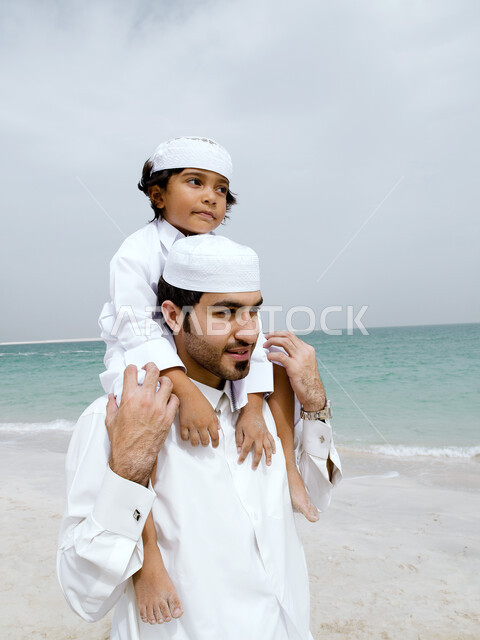 الإستمتاع بالمشي على الشاطئ في يوم مشمس, قضاء وقت ممتع مع الابناء, أب إماراتي عربي خليجي يرتدي الكندورة والغترة الإماراتيه يحمل ابنه على كتفيه, رحله عائليه سعيدة في سواحل الإمارات, إجازه صيفيه ممتعة, نشاطات ترفيهية مسلية، إيماءات وجه تدل على السعادة 