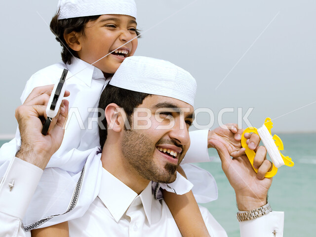 الإستمتاع باللعب في يوم مشمس, قضاء وقت ممتع مع الابناء, أب إماراتي عربي خليجي يلعب مع ولده بالطائرة الورقية ويضعه على كتفيه, رحله عائليه سعيدة في سواحل الإمارات, إجازه صيفيه ممتعة, نشاطات ترفيهية مسلية، إيماءات وجه تدل على السعادة 