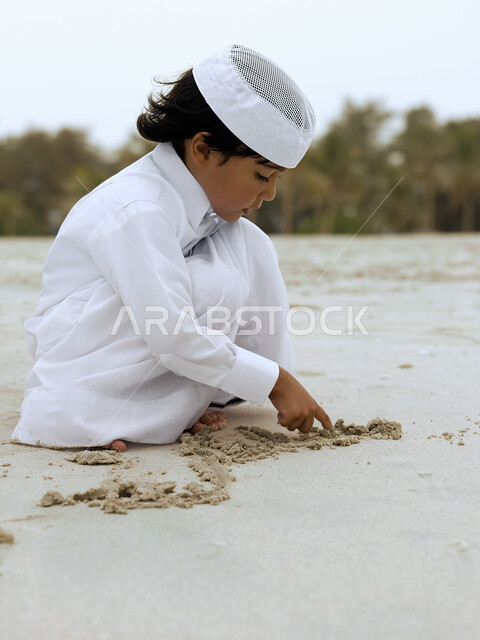 الاستمتاع بالعطلة الصيفيه, فتى عربي إماراتي خليجي يرتدي الزي الإماراتي التقليدي يلعب على شاطيء البحر بالرمال الذهبية الناعمة، نشاطات ترفيهية مسلية وقضاء أوقات ممتعة, السياحه في الإمارات العربية المتحدة، خلفية الطبيعة.