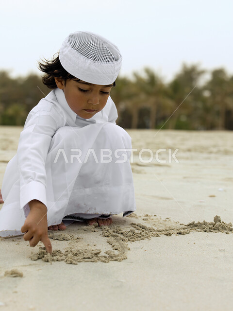 الاستمتاع بالعطلة الصيفيه, فتى عربي إماراتي خليجي يرتدي الزي الإماراتي التقليدي يلعب على شاطيء البحر بالرمال الذهبية الناعمة، نشاطات ترفيهية مسلية وقضاء أوقات ممتعة, السياحه في الإمارات العربية المتحدة، خلفية الطبيعة.