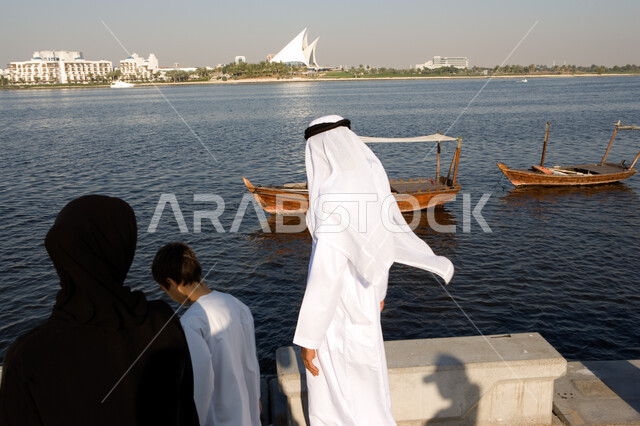 الاستمتاع بمشاهدة القوارب الخشبية في البحر, مفهوم الترابط الأسري وقضاء الوقت بالسير مع الأبناء , الاهتمام بالعائلة و مشاركتهم اللحظات السعيدة, صورة من الخلف لعائلة عربية خليجية إماراتية تقوم بالتنزه والمشي على الكورنيش في يوم العطلة, الحوارات والنقاشات ال