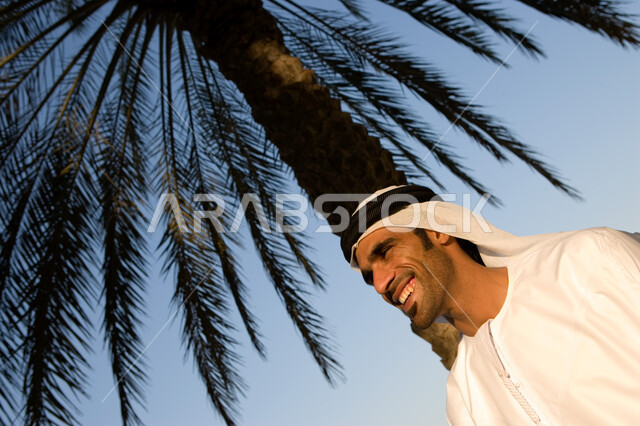 الاحتماء من حر الشمس تحت ظلال شجرة النخيل, صورة مقربة لرجل عربي خليجي إماراتي يرتدي الغترة والكندورة الإماراتية يقف على الكورنيش تحت شجرة النخيل بإيماءات تدل على السعادة, قضاء وقت الإجازة بالاستمتاع بالهواء الطلق, السياحة في الامارات العربية المتحدة